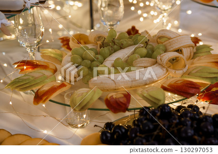 Festive Fruit Platter Arrangement with Bananas, Grapes, Apples and Cherries Illuminated by Warm String Lights, White Tablecloth Setting 130297053