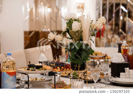 Elegant Dinner Table Setting with Pampas Grass Arrangement, White Floral Centerpiece, Crystal Glassware, and Appetizer Platters in a Luxury Interior Elegant Dinner Table Setting with Pampas Grass Arrangement, White Floral Centerpiece, Crystal Glassware, and Appetizer Platters in a Luxury Interior 130297105