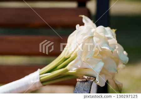 Wedding Rings and White Calla Lilies Bouquet on Wooden Bench. Love, Commitment, Marriage Symbolism, Close-Up, Outdoor Scene, Soft Focus, Natural Light. Wedding Rings and White Calla Lilies Bouquet on Wooden Bench. Love, Commitment, Marriage Symbolism, Close-Up, Outdoor Scene, Soft Focus, Natural Light. 130297125