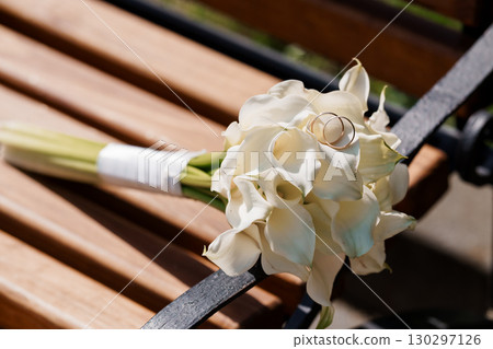 Elegant Wedding Rings on White Calla Lily Bouquet, Rustic Wooden Bench, Close-Up, Romantic Celebration, Sunlit Still Life 130297126