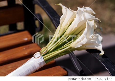 Elegant Wedding Bouquet of White Calla Lilies and Gold Wedding Rings on Wooden Park Bench, Romantic Celebration Style. Elegant Wedding Bouquet of White Calla Lilies and Gold Wedding Rings on Wooden Park Bench, Romantic Celebration Style. 130297128