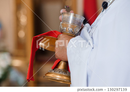 Priest Holding Silver Chalice During Religious Ceremony. Red Cloth. Orthodox Church Interior. Faith, Spirituality, Tradition. Priest Holding Silver Chalice During Religious Ceremony. Red Cloth. Orthodox Church Interior. Faith, Spirituality, Tradition. 130297139
