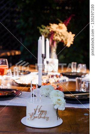 Intimate Birthday Celebration Scene with White Cake, Candles, Floral Arrangement and Warm Lighting in a Rustic Outdoor Setting. 130297180