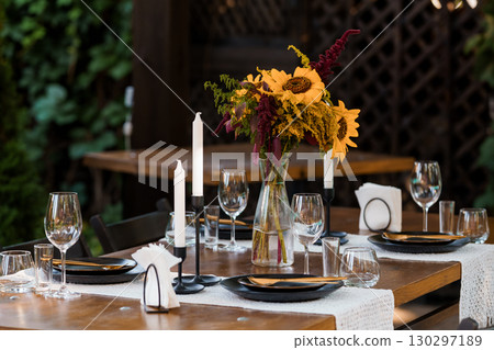 Rustic Outdoor Dinner Setting with Sunflowers and Candles: Warm Autumn Table Decor for Cozy Events, Dark Plates, and Wooden Texture. Rustic Outdoor Dinner Setting with Sunflowers and Candles: Warm Autumn Table Decor for Cozy Events, Dark Plates, and Wooden Texture. 130297189
