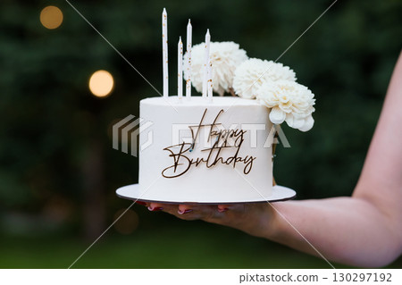 Elegant Birthday Cake with White Flowers and Candles Held by Hand, Creamy White Frosting, Celebration, Party Decor, Outdoor Setting 130297192