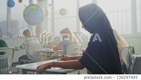 Young Muslim Girl Wearing Hijab Sitting at the Desk, Writing School Exam in Notebook 130297485