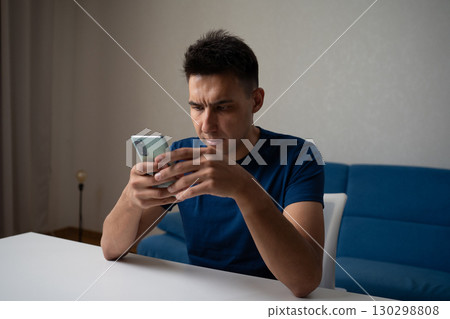 Young adult man in casual clothes using mobile phone sitting at table, browsing internet or social media, concentrating on online content at home. Concept of modern communication and technology. Young adult man in casual clothes using mobile phone sitting at table, browsing internet or social media, concentrating on online content at home. Concept of modern communication and technology. 130298808