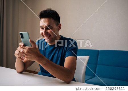 Cheerful young man in casual clothes using mobile phone sitting at table, browsing internet or social media, smiling on online content at home. Concept of modern communication, technology. Cheerful young man in casual clothes using mobile phone sitting at table, browsing internet or social media, smiling on online content at home. Concept of modern communication, technology. 130298811
