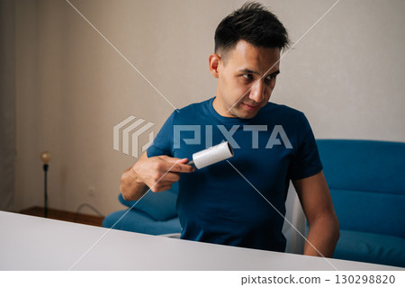 Young man sitting at white table using sticky lint roller to remove dust, hair and lint from blue t-shirt, looking pleased with cleaning results in modern apartment. Concept of household cleanliness. Young man sitting at white table using sticky lint roller to remove dust, hair and lint from blue t-shirt, looking pleased with cleaning results in modern apartment. Concept of household cleanliness. 130298820