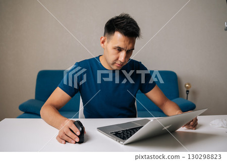 Ufa, Russia - 30 July 2025. Portrait of freelancer male concentrating on work sitting at table in modern home office, using vertical mouse and laptop to enhance productivity and maintain focus. Ufa, Russia - 30 July 2025. Portrait of freelancer male concentrating on work sitting at table in modern home office, using vertical mouse and laptop to enhance productivity and maintain focus. 130298823