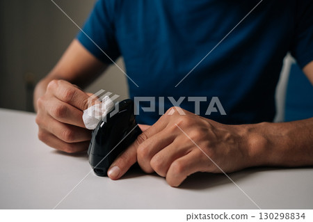 Close-up cropped shot of man cleaning modern black vertical mouse with disinfectant wipe, promoting hygiene and preventing spread of germs, sitting at white table in workplace or home office. 130298834
