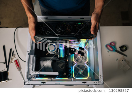 Top view of computer technician installing high-end graphic card into custom gaming pc, showcasing vibrant rgb lighting and advanced water cooling for peak performance enhancement. 130298837