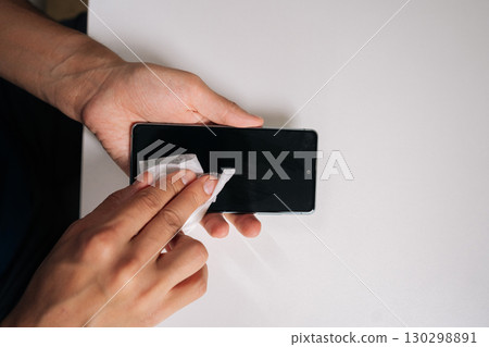 High-angle view of man carefully wiping smartphone screen with disinfectant wipe, ensuring hygiene and removing germs and bacteria from device display. Concept of cleanliness and housekeeping. 130298891