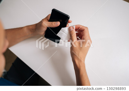 View from shoulder of man cleaning screen of smartphone with disinfectant wipe, promoting hygiene and effectively eliminating germs and bacteria for better health, sitting at white desk View from shoulder of man cleaning screen of smartphone with disinfectant wipe, promoting hygiene and effectively eliminating germs and bacteria for better health, sitting at white desk 130298893