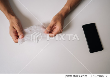 Top view of unrecognizable man preparing to cleaning smartphone screen using wet wipe and alcohol pad, ensuring hygiene and removing dirt and fingerprints from device surface sitting at desk at home. 130298897