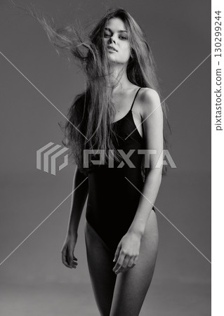 Black and white portrait of young woman in black bodysuit with flowing hair and serious expression on gray background 130299244