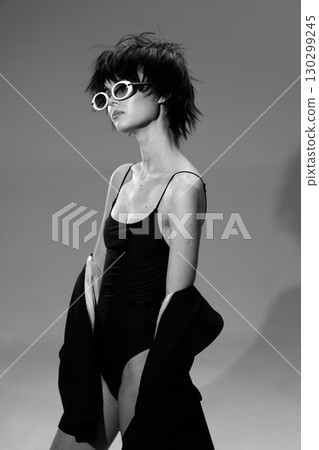 Black and white portrait of a stylish woman wearing sunglasses and a black dress with jacket, posing confidently indoors. Black and white portrait of a stylish woman wearing sunglasses and a black dress with jacket, posing confidently indoors. 130299245