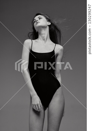 Black and white portrait of a slender woman in a black leotard posing with a serene expression on a plain gray background. 130299247