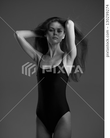 Black and white portrait of a slender woman in a black swimsuit posing confidently against a gray background. Black and white portrait of a slender woman in a black swimsuit posing confidently against a gray background. 130299274
