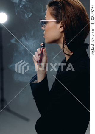 Stylish woman vaping in dark indoor setting with smoke and modern lighting, wearing glasses and black jacket. Stylish woman vaping in dark indoor setting with smoke and modern lighting, wearing glasses and black jacket. 130299275