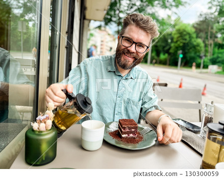Man pouring tea into cup while smiling at outdoor cafe table. Social lifestyle moment of relaxation, food and drink enjoyment in urban setting. 130300059