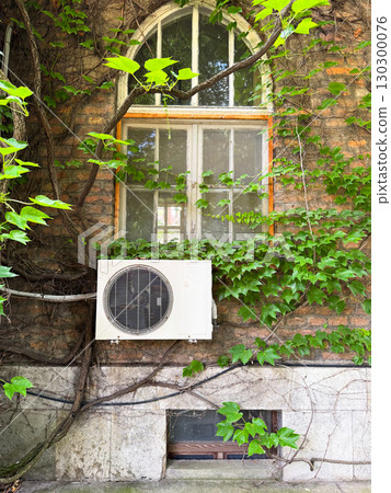 Arched window with ivy and air conditioning unit on brick wall. Cultural architecture combined with modern technology and natural greenery in urban environment. 130300076