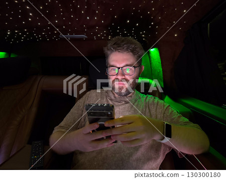 Man focused on smartphone inside luxury illuminated vehicle. Concentration, technology and modern lifestyle in travel environment. 130300180