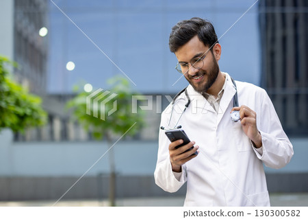 Young male medical doctor smiling while using a mobile phone app and holding a stethoscope, enjoying a moment outdoors in front of the hospital on a sunny day Young male medical doctor smiling while using a mobile phone app and holding a stethoscope, enjoying a moment outdoors in front of the hospital on a sunny day 130300582