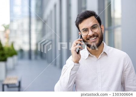 Young businessman is having a pleasant conversation on his mobile phone while standing outside a modern office building, enjoying the urban environment Young businessman is having a pleasant conversation on his mobile phone while standing outside a modern office building, enjoying the urban environment 130300604