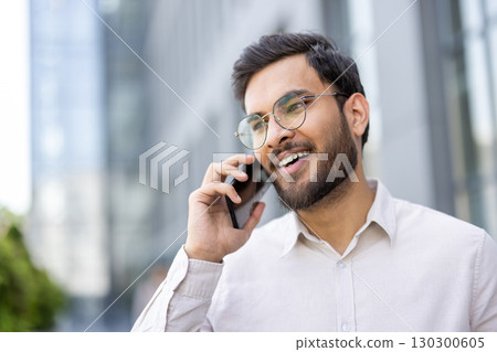 Portrait of a smiling young businessman wearing glasses, speaking on his mobile phone in front of an office building, enjoying a pleasant conversation Portrait of a smiling young businessman wearing glasses, speaking on his mobile phone in front of an office building, enjoying a pleasant conversation 130300605