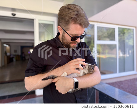 Kitten resting calmly in arms of man with beard outdoors. Trust, safety, and emotional comfort through human animal interaction. Kitten resting calmly in arms of man with beard outdoors. Trust, safety, and emotional comfort through human animal interaction. 130300611