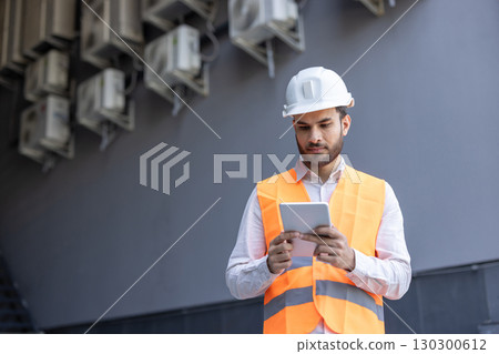 Building maintenance technician wearing safety helmet and vest, using digital tablet and inspecting air conditioning units on building exterior Building maintenance technician wearing safety helmet and vest, using digital tablet and inspecting air conditioning units on building exterior 130300612
