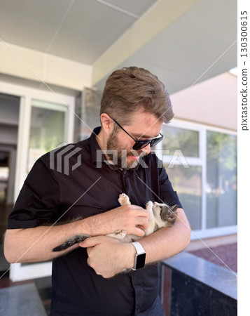 Bearded man gently holding kitten in hands and looking down. Care, compassion and emotional connection with domestic animal. 130300615