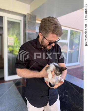 Man in sunglasses smiling and holding small kitten outdoors. Companionship affection, and emotional bond between human and animal. 130300617