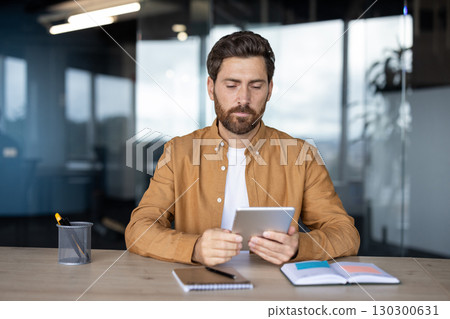 A professional man using a digital tablet while seated at a desk in a modern office. The environment conveys focus, productivity, and technology in a corporate workspace. A professional man using a digital tablet while seated at a desk in a modern office. The environment conveys focus, productivity, and technology in a corporate workspace. 130300631