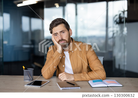 A man sits at a desk in an office, looking bored and resting his head on his hand, surrounded by office supplies. A man sits at a desk in an office, looking bored and resting his head on his hand, surrounded by office supplies. 130300632