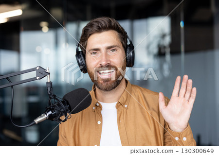 A man with a beard waves while wearing headphones and recording a podcast. He smiles near a microphone in a studio setting. 130300649