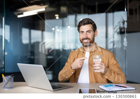 Happy businessman promoting healthy lifestyle, sitting at desk with laptop and pointing at glass of water, smiling at camera in modern office workspace 130300661
