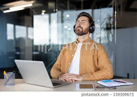 Office worker enjoying music with closed eyes, taking a well-deserved break from tasks at his desk, surrounded by a laptop, tablet, and notebook, embracing relaxation and tranquility 130300673