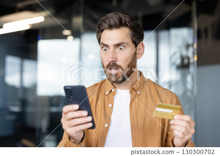 A shocked man looks at his phone screen while holding a credit card, possibly reacting to an unexpected charge or financial issue. A shocked man looks at his phone screen while holding a credit card, possibly reacting to an unexpected charge or financial issue. 130300702