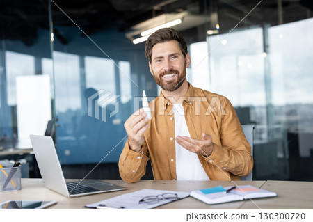 Man sitting at a modern office desk, presenting a small bottle of nose drops with a smile. 130300720