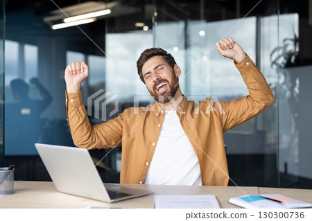 A happy man celebrates a win at his office desk, laptop open, hands raised in excitement and joy. 130300736