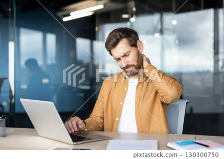 A man in an office setting experiencing neck pain, sitting at a desk with a laptop. The image highlights workplace discomfort and injury awareness in a corporate working environment. 130300750