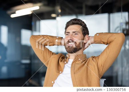Smiling businessman is sitting relaxed in his chair with his hands behind his head, enjoying a moment of peace and quiet in the modern office 130300756