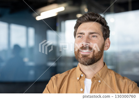 Smiling man wearing a casual shirt standing in a bright contemporary office space with large windows. The professional setting and his positive expression reflect confidence, success 130300760