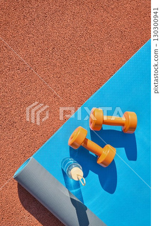 Reusable glass bottle of water, dumbbells and blue yoga mat at stadium 130300941