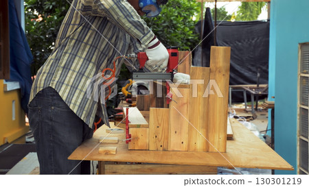 Carpenter using pneumatic nail gun to secure wooden plank onto vertical timber assembly at outdoor workshop 130301219
