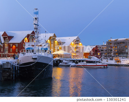 Evening scene at Tromso harbor featuring docked boats under a snowy landscape. Warm lights from red and yellow buildings reflect on the water, creating a serene winter atmosphere Evening scene at Tromso harbor featuring docked boats under a snowy landscape. Warm lights from red and yellow buildings reflect on the water, creating a serene winter atmosphere 130301241