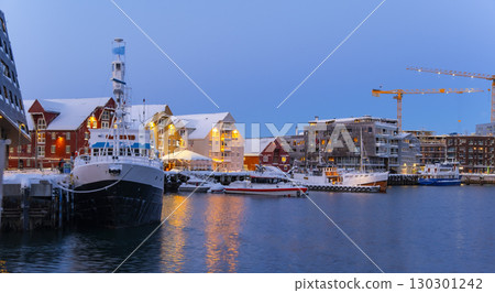 Evening scene at Tromso harbor featuring docked boats under a snowy landscape. Warm lights from red and yellow buildings reflect on the water, creating a serene winter atmosphere 130301242
