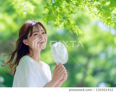 一位微笑的日本女士拿著小電風扇享受清涼，周圍是清新的綠植和清爽的夏日景色 130301432